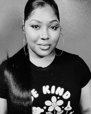 A black-and-white portrait of a woman with long hair pulled back, wearing large hoop earrings and a T-shirt that reads ‘Be Kind’. She is looking directly at the camera with a calm expression