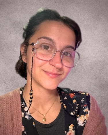 A woman smiling softly at the camera, wearing large round glasses with a beaded glasses chain, a floral patterned top layered with a pink cardigan, and a gold heart necklace. She is posed in front of a textured gray background.