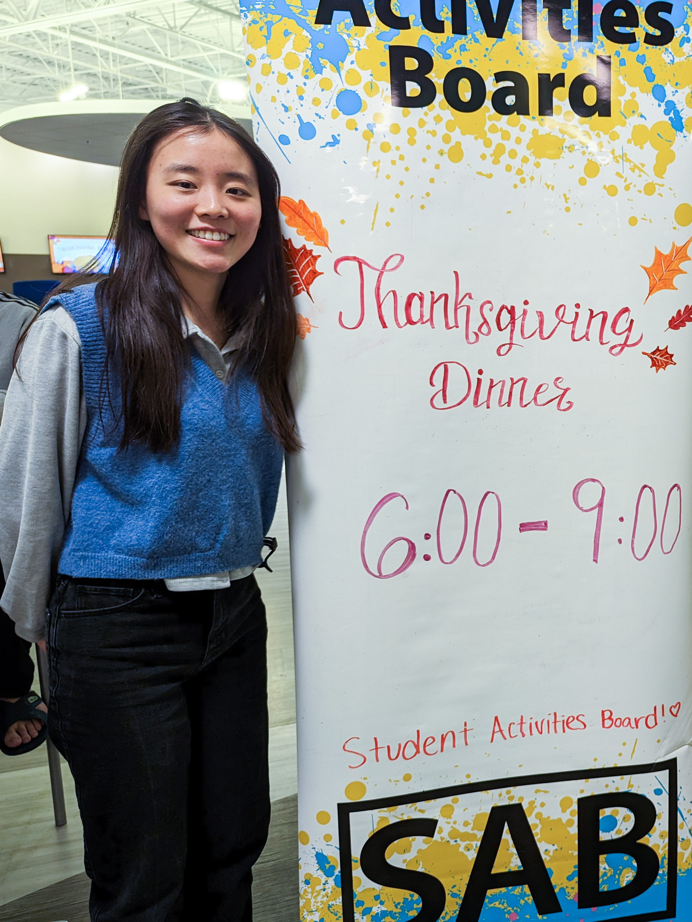 Sophomore Eejoy Lim poses near an event sign for the 2022 UM-Dearborn Friendsgiving