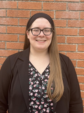 Head-and-shoulders portrait of a smiling person standing in front of a brick wall, wearing glasses, a black headband, a dark blazer, and a black floral blouse, with long hair draped over one shoulder.
