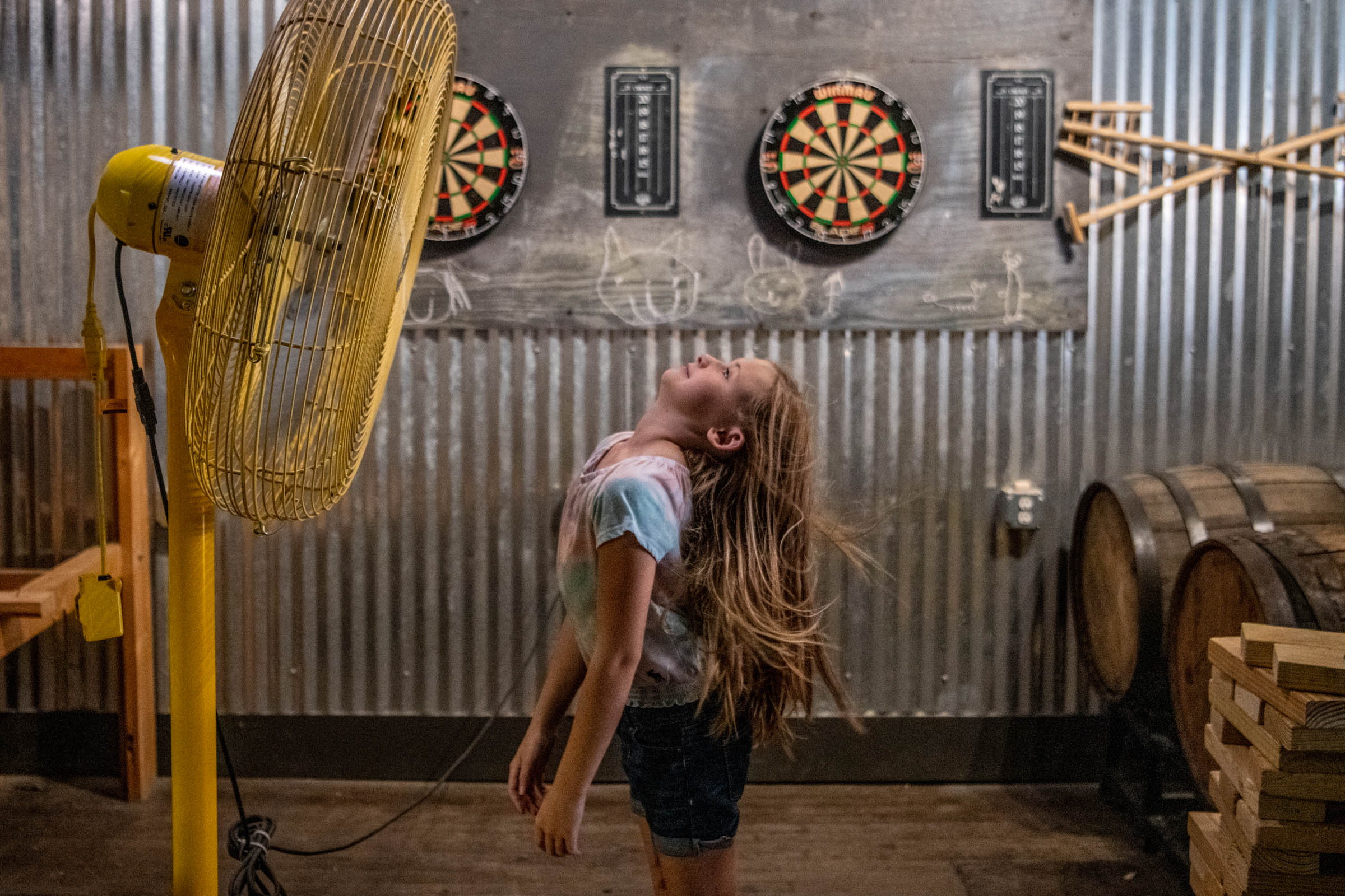 In a warehouse backroom, a young girl stands in front of a large fan, the breeze blowing her head and hair back