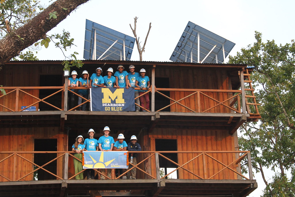 Students wearing white hard hats and turquoise t-shirts pose for a photo on a wooden watchtower with a UM-Dearborn banner draped over the railing.