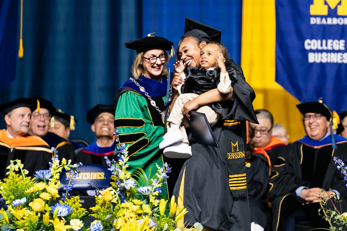 Two women and a toddler gather on the graduation stage.  The background is decorated with maize and blue banners.