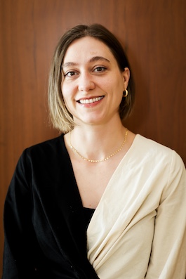 a smiling person with short blonde/brown hair poses in front of a wooden backdrop wearing a black and white wrap top with gold jewelry