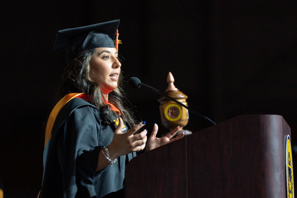 A female graduate, who is wearing a cap and gown, speaks at a commencement ceremony. She is at a podium and talking into a microphone.