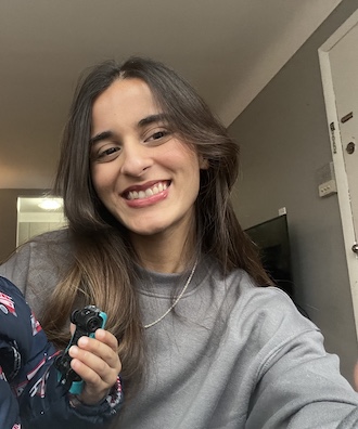 Smiling person taking a selfie indoors, wearing a gray sweatshirt, with long dark hair and a TV and doorway visible in the background