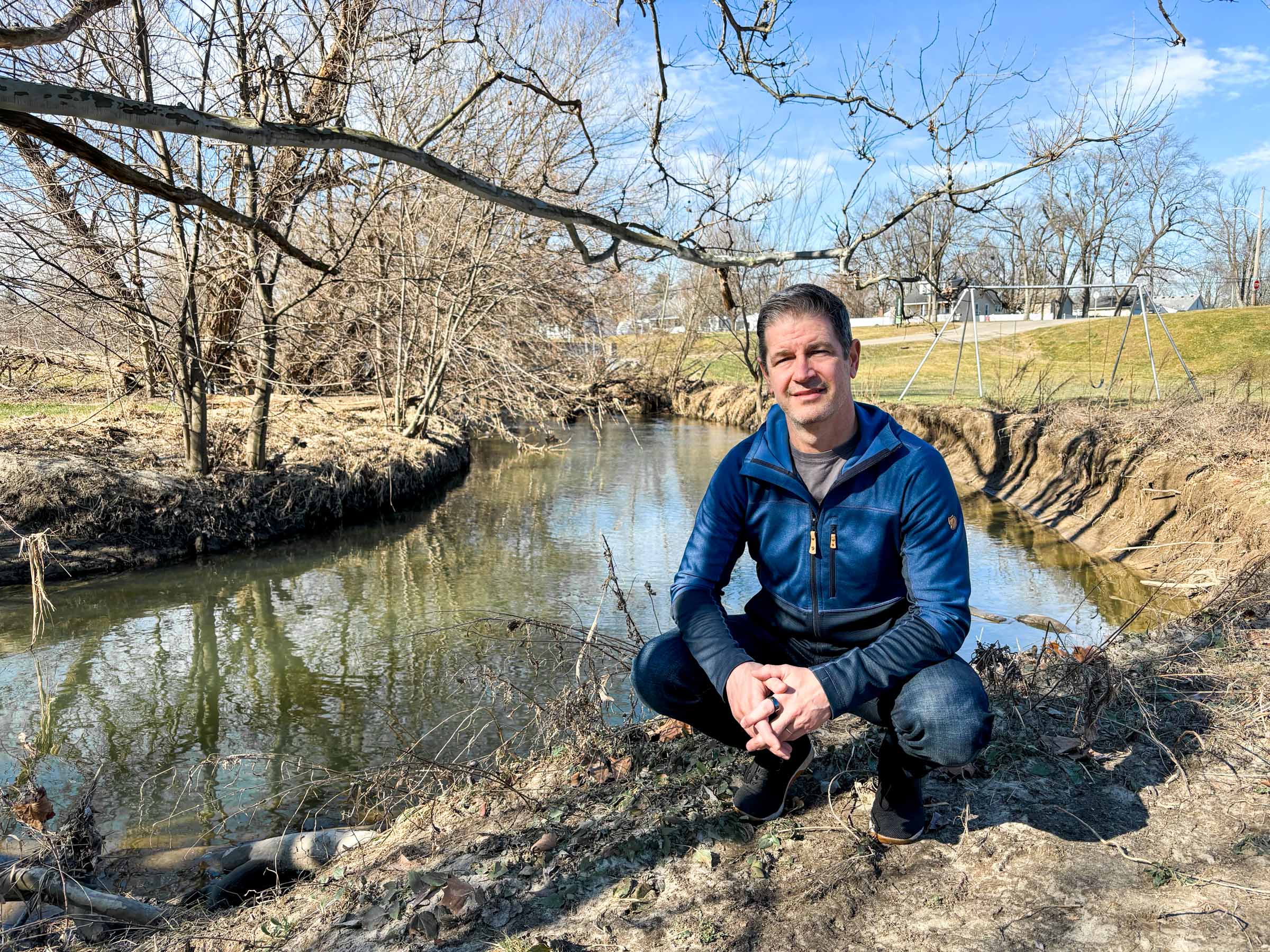 Professor Jacob Napieralski squats next to the Rouge River on a late winter day