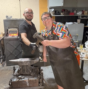 Two people in aprons smiling while holding a hammer together in a blacksmith workshop with an anvil and forge.