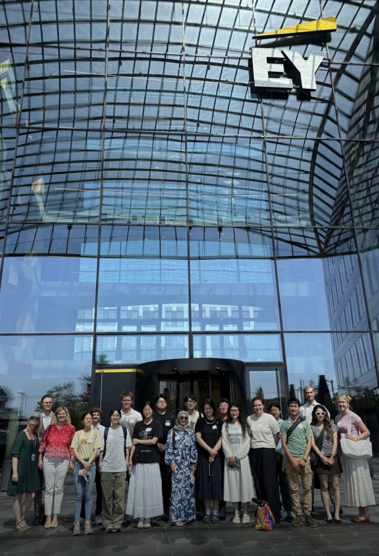 Group of students and faculty posing for a photo outside an EY office building, beneath a large glass atrium and the EY logo.