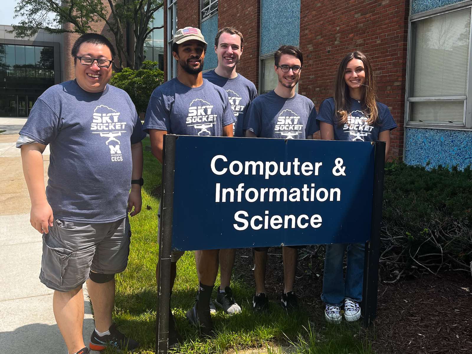 Wearing their custom "Sky Socket" project t-shirts, Assistant Professor Zheng Song stands with student Olivia Pellegrini and her senior design teammates outside the nameplate for the Computer and Information Science Building on a summer day. 