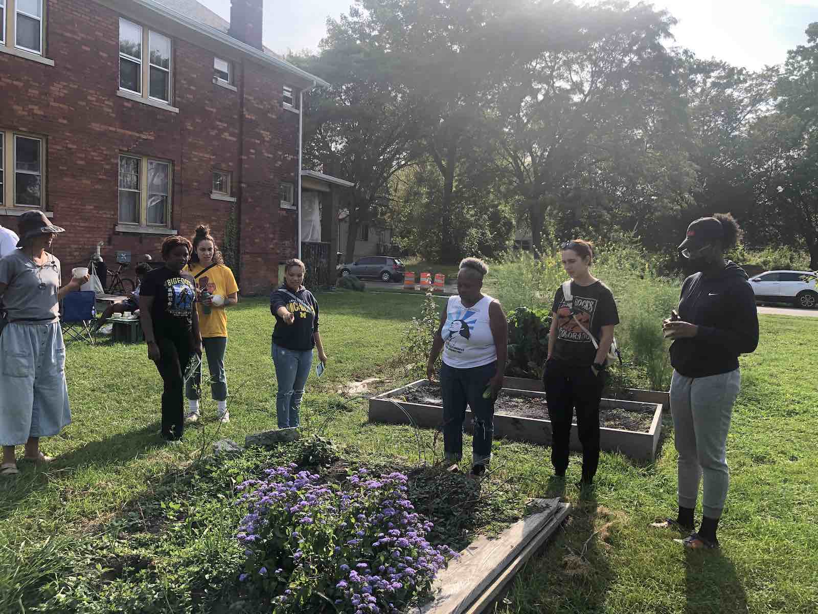 College students and a community member check out a community garden in an open lot next to a brick house in a Detroit neighborhood