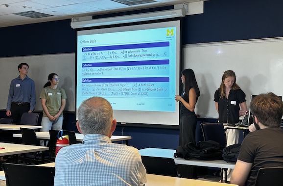 A group of students presents mathematical definitions on a projector screen in a classroom.Audience members listen attentively and take notes at tables.The University of Michigan logo is displayed on the presentation slide.