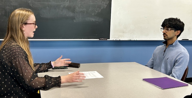 Two people are seated across from each other at a table, engaged in conversation.There are documents and a purple folder on the table, suggesting an interview or meeting. A chalkboard and whiteboard are visible in the classroom background.
