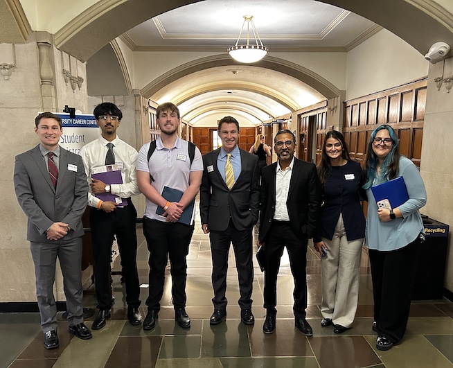A group of professionally dressed individuals stands together in a university hallway.   They wear nametags and hold folders, indicating attendance at a career event.   A sign for the Student Registration area is visible in the background.