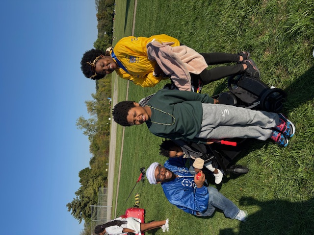 Three people stand together outdoors on a sunny day, with a stroller beside them. One person in a yellow jacket stands behind a child wearing a green hoodie, while another person in a blue jacket and white headwrap kneels next to the stroller. They are gathered on a grassy field with trees in the background