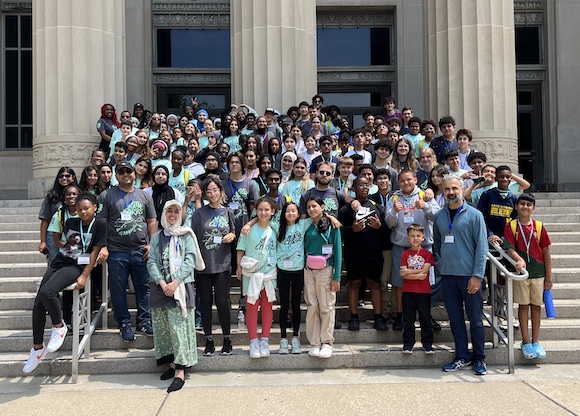 A large group poses on stone steps in front of a classical building with tall columns.Most participants wear Math Corps shirts and lanyards, suggesting a camp or event.The scene is bright and energetic, capturing a moment of togetherness and community.
