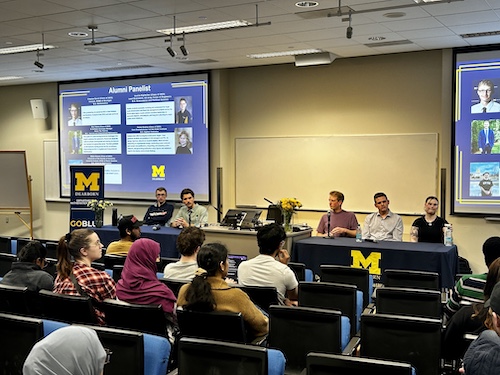 Panel of alumni speaking to students in a university classroom.Presentation slide titled "Alumni Panelist" shown on projector.University of Michigan Dearborn banners and logo visible in the room.