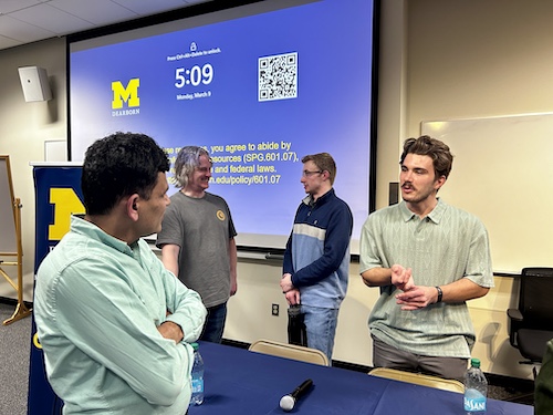 Three people stand and converse in front of a University of Michigan Dearborn presentation screen.A blue and yellow UM-Dearborn banner is positioned beside them.The screen displays a QR code, the time, and campus policy information.