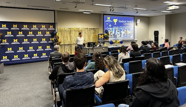 Audience seated in a lecture hall facing a podium and presentation screen.Slide reads “Student Award Ceremony” with University of Michigan–Dearborn branding.Maize-and-blue balloons, gold streamers, and a U-M step-and-repeat backdrop decorate the front.