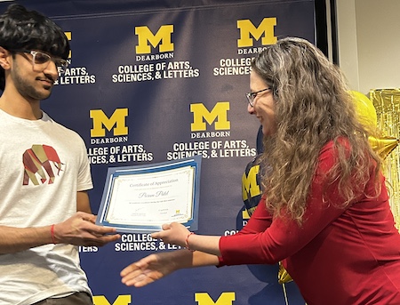 Two people exchange a Certificate of Appreciation in front of a UM–Dearborn CASL backdrop.The recipient holds the framed certificate while the presenter reaches toward it.Maize-and-blue balloons and gold decorations appear at the right edge.