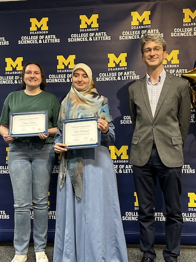 Three people pose in front of a UM–Dearborn College of Arts, Sciences, & Letters backdrop.Two hold Certificate of Appreciation awards while the third stands beside them.A gold balloon is partially visible at the right edge.
