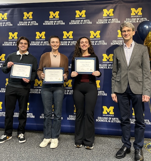 Four people pose in front of a UM–Dearborn College of Arts, Sciences, & Letters backdrop.Three hold Certificate of Appreciation awards while one stands at the right.A cluster of maize-and-blue balloons is visible along the right edge.