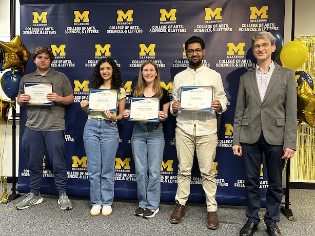 Five people pose in front of a UM–Dearborn College of Arts, Sciences, & Letters backdrop.Four hold certificates while a fifth stands at the right in a blazer.Maize-and-blue balloons and gold streamers frame the scene at both edges.