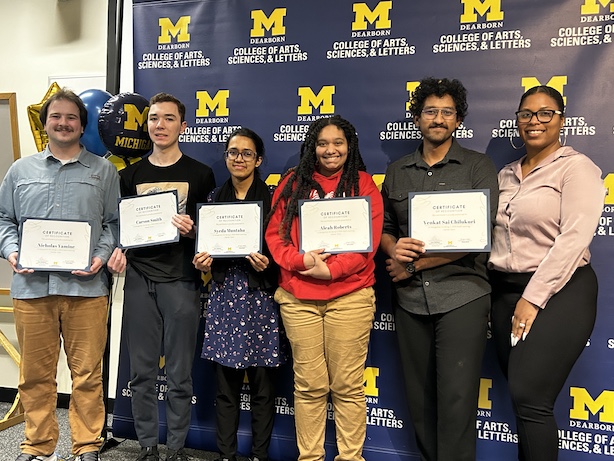Six people pose in front of a UM–Dearborn College of Arts, Sciences, & Letters backdrop.Five hold certificates while one stands at the far right.Maize-and-blue balloons are visible at the left edge.