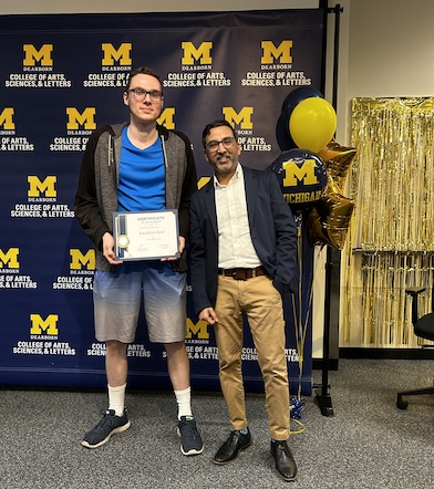 Two people pose in front of a University of Michigan–Dearborn College of Arts, Sciences, & Letters backdrop.One holds a certificate while the other stands beside them with hands in pockets.Maize-and-blue balloons and gold streamers decorate the area to the right.
