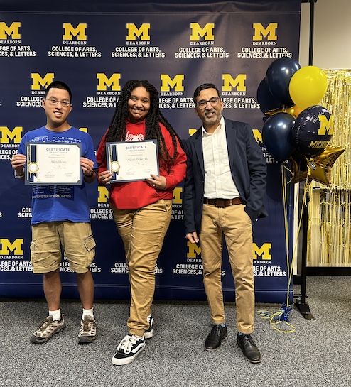 Three people pose in front of a UM–Dearborn College of Arts, Sciences, & Letters backdrop.Two hold certificates while a third stands beside them in business attire.Maize-and-blue balloons and gold streamers decorate the right side.