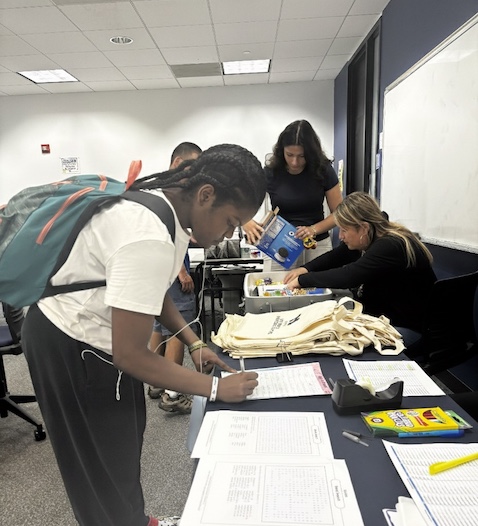 Student with backpack signing her name on a paper with 3 people in the background filing documents.