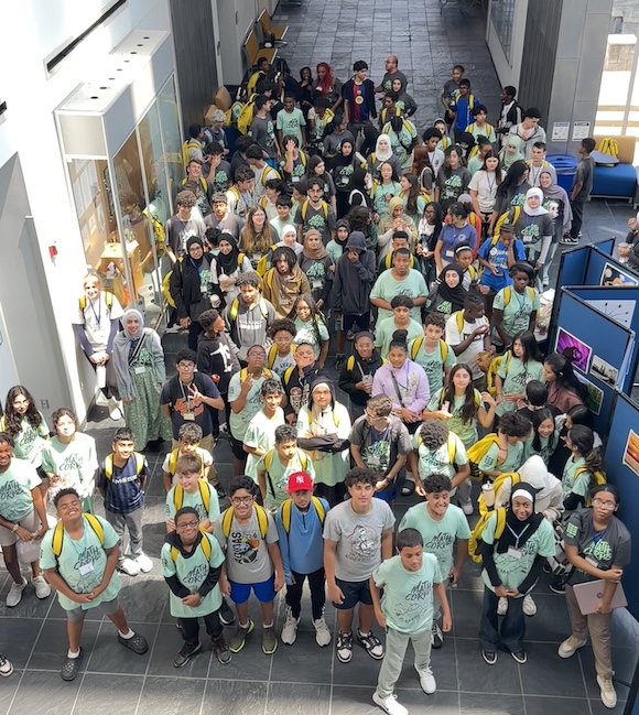 A large group of students stands together in a bright indoor hallway, many wearing Math Corps shirts and backpacks. Some students are looking up, grouped closely in an open, spacious area with natural light. Artwork is displayed on the right side, and yellow backpacks are visible throughout the crowd.