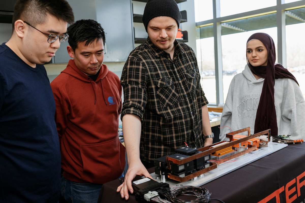 Four students gather around a laboratory table, focused on a complex electronic assembly with copper components and wiring. A man wearing a black beanie and plaid shirt points down toward a control module while three teammates observe the engineering project.
