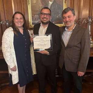 3 people pose with the center person holding a certifcate award, the left wears a dark dress with a cream sweater, the right wears a brown blazer and dark pants, and the middle is in a dark suit