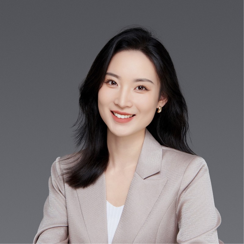 A woman with shoulder-length black hair smiles warmly at the camera. She wears small gold earrings and a light beige blazer over a white top against a plain gray background.
