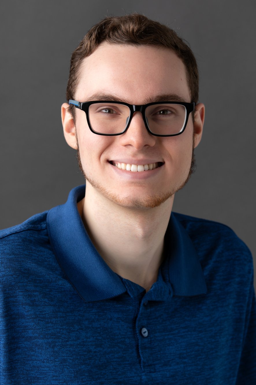 Head-and-shoulders studio portrait of a person in a blue collared polo shirt.Face is blurred/obscured, with short brown hair visible.Neutral dark gray background with soft, even lighting.