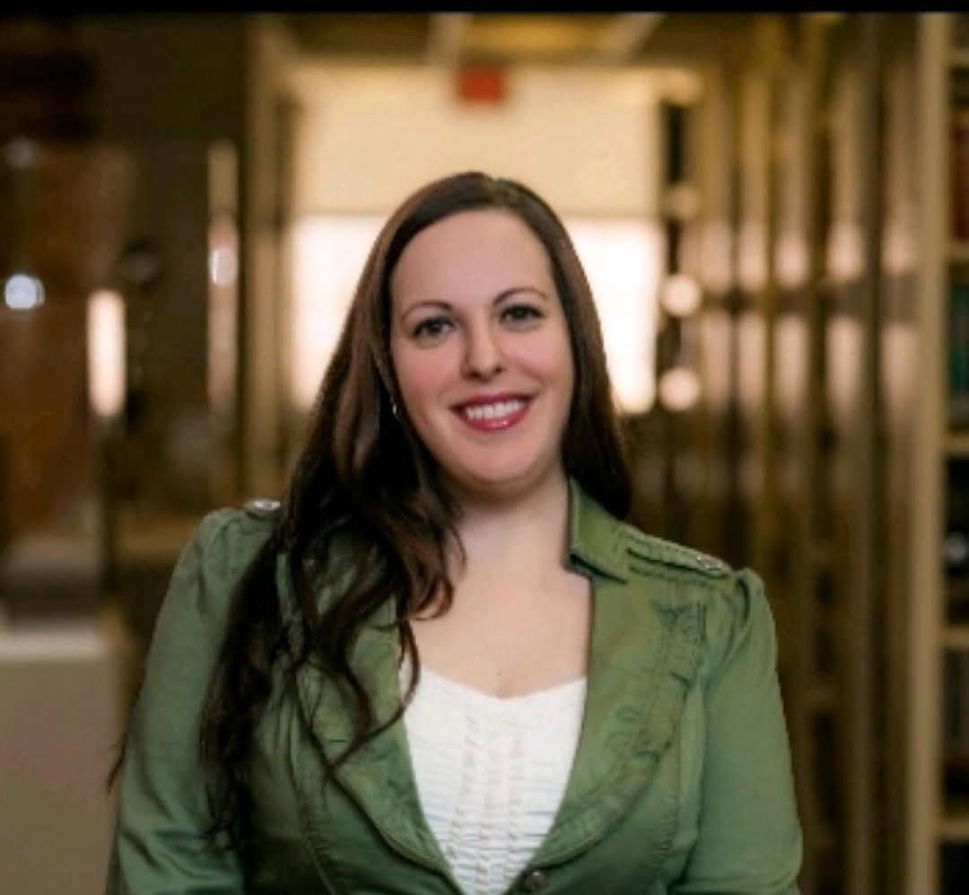 a smiling person with long dark hair wearing a green jacket and cream top stands in front of a hallway and book cases