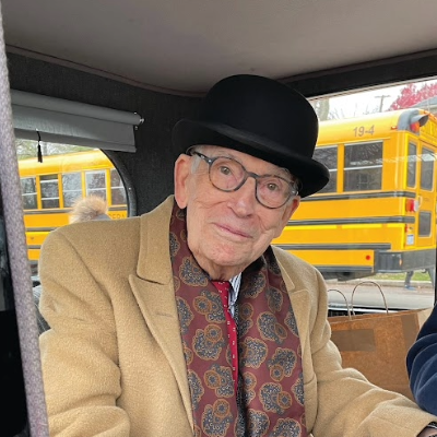 Judge Van Tiem wearing glasses, a tan coat, a patterned scarf, and a black bowler hat smiles while sitting inside a vehicle, with yellow school buses visible through the window behind him.