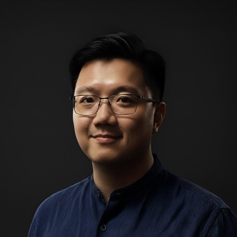A man with short, neatly styled dark hair and rectangular glasses looks at the camera with a slight smile. He wears a dark blue collarless shirt against a black background.