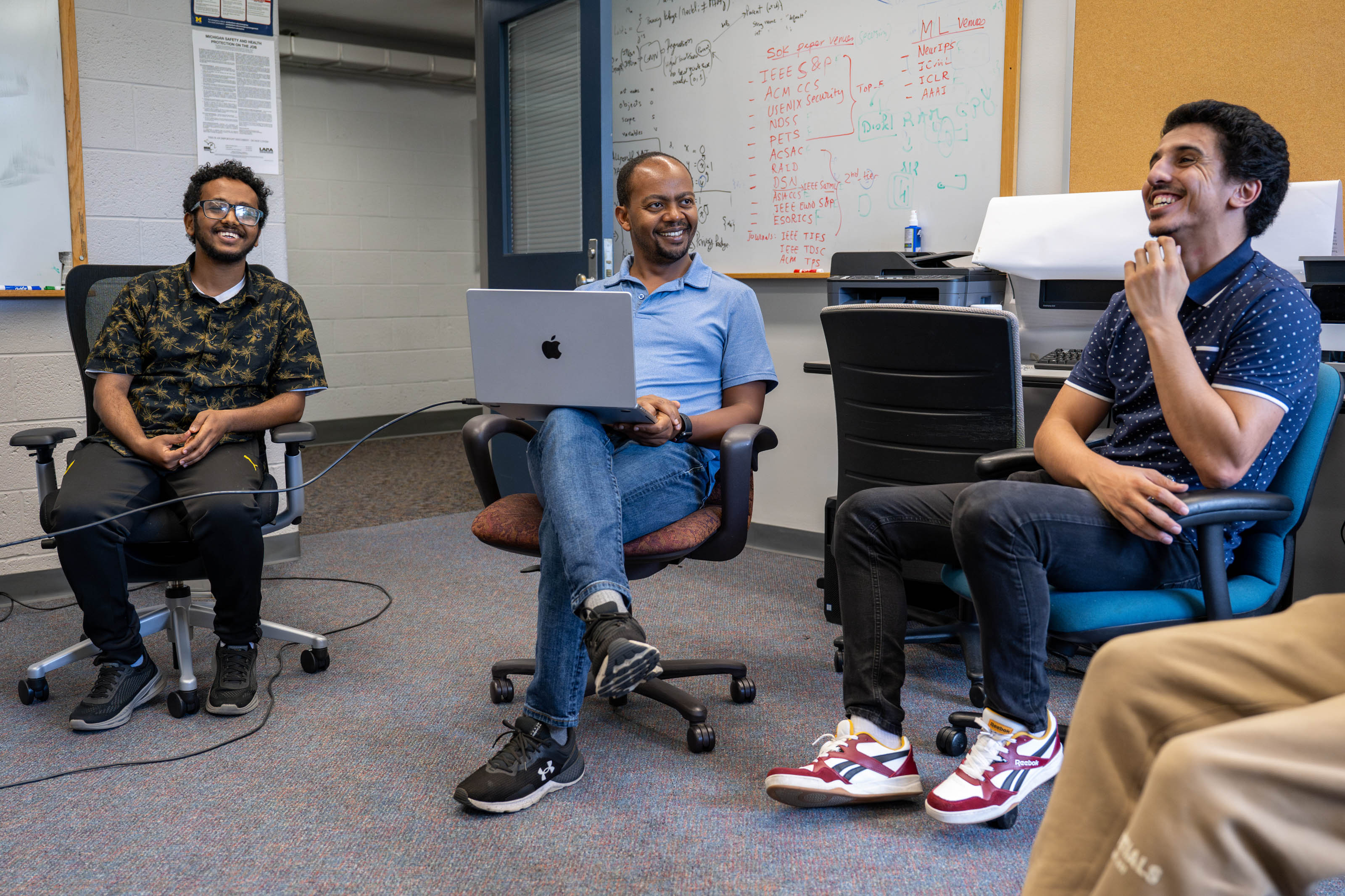 Associate Professor Birhanu Eshete sitting and laughing with two of his graduate students in their artificial intelligence lab.