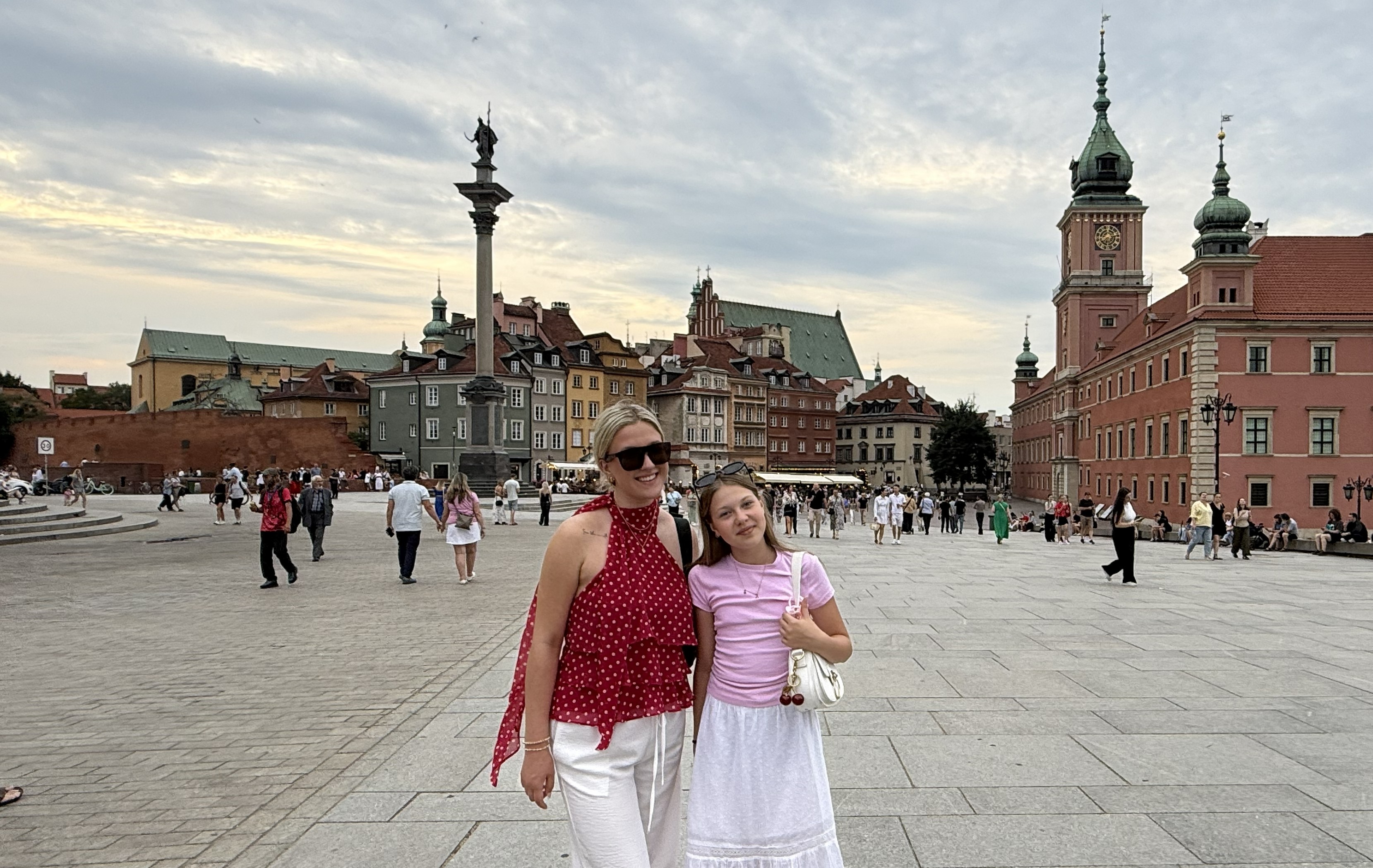 Kaelyn Mlostek poses with another woman in Warsaw's Old Town square in Poland, with historic colorful buildings, Sigismund's Column and the Royal Castle in the background.