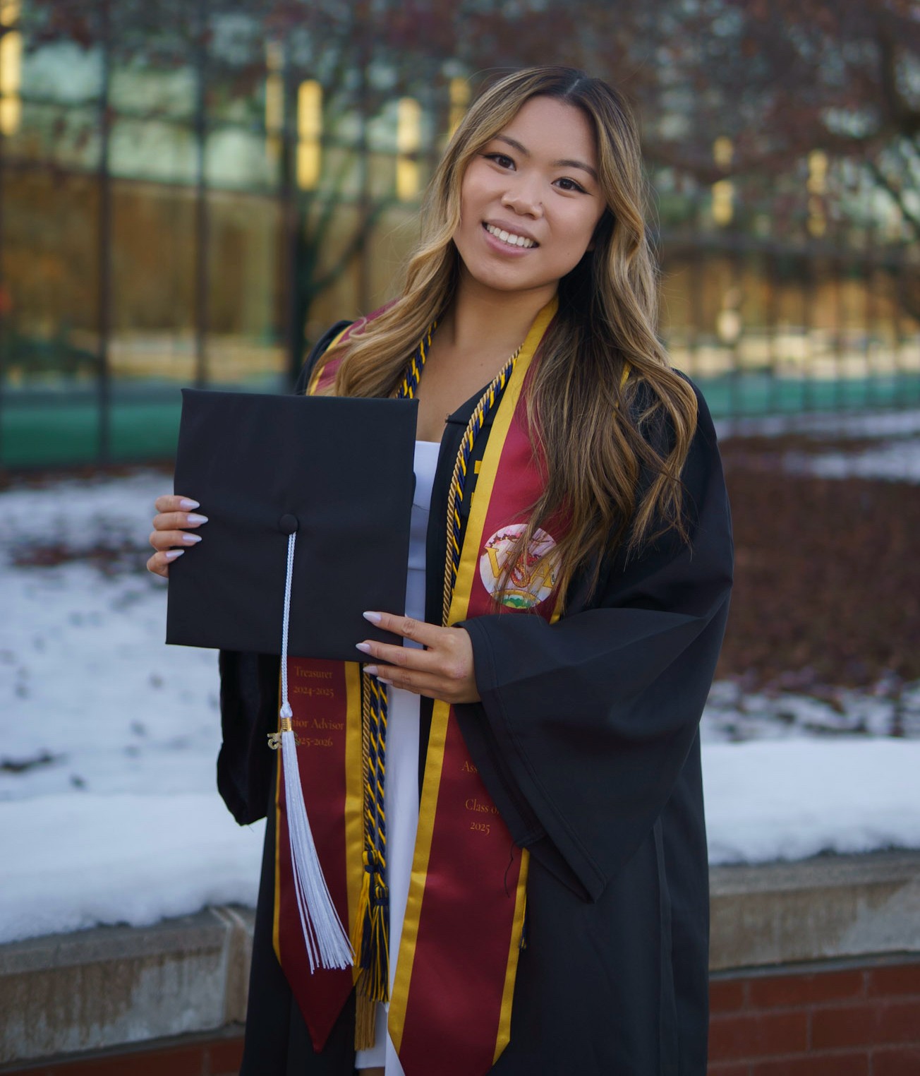 Graduate in a black gown and red-and-gold stole holds a graduation cap with a white tassel.Long hair falls over the shoulders; a white outfit is visible beneath the gown.Snowy ground and a glass building appear in the blurred background.