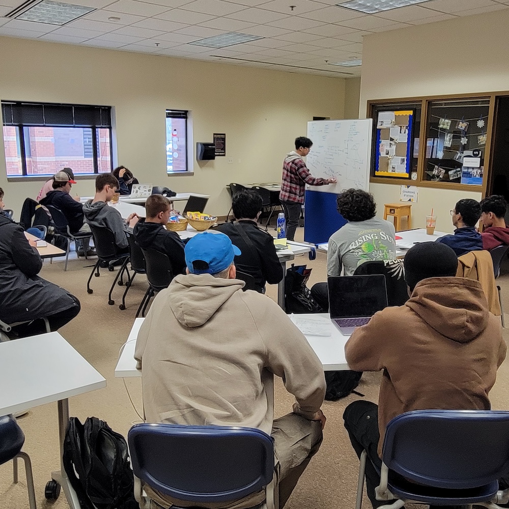 A classroom setting with students seated at desks using laptops, focusing on a presentation being given by a person at the front beside a whiteboard.
