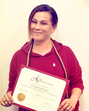 A woman smiling softly while holding an Alpha Sigma Lambda certificate of membership. She is wearing a red textured hoodie and an honor cord around her neck