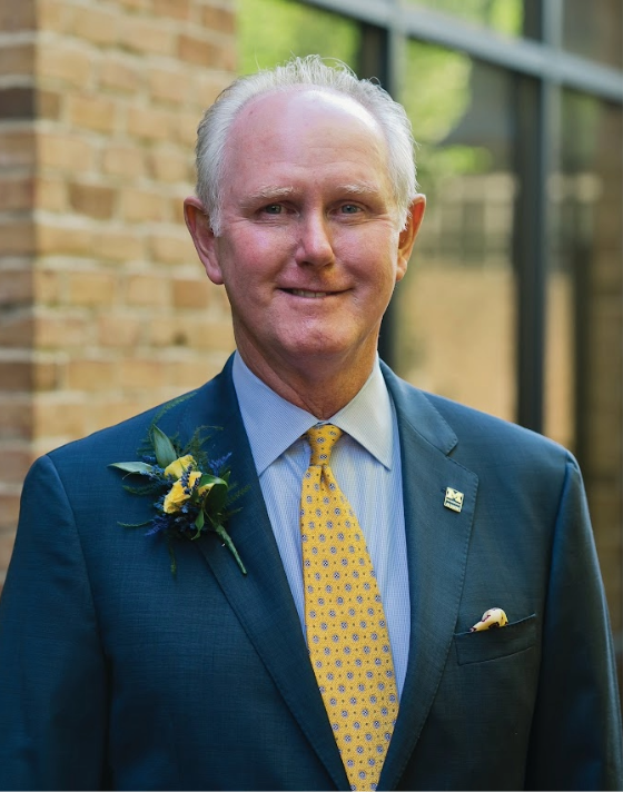 Kris Pfaehler wearing a navy blue suit, light blue shirt, and yellow patterned tie stands outdoors beside a brick building. He has a boutonniere pinned to his jacket and smiles directly at the camera in soft, natural light.