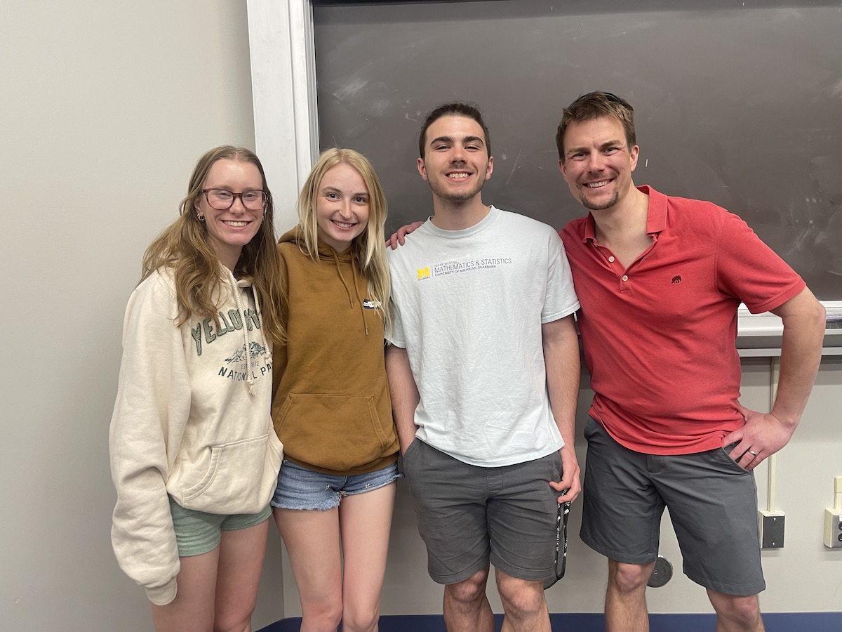 Four people stand closely together in a classroom in front of a chalkboard.They are casually dressed in shorts and t-shirts or hoodies.The atmosphere appears relaxed and friendly, suggesting a group project or study session.