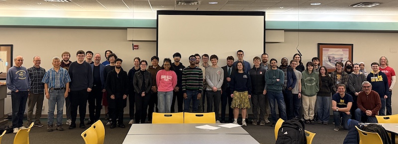 Large group of people pose in a classroom or meeting room in front of a projector screen.Several wear University of Michigan apparel; tables and yellow chairs are in the foreground.Two people kneel at the right while the rest stand in a single line.