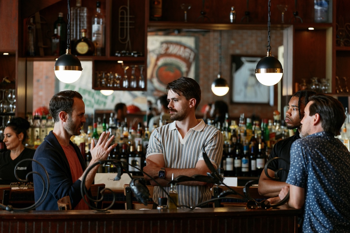 Four males talk to one another with an antique wooden bar with a centered mirror in the background