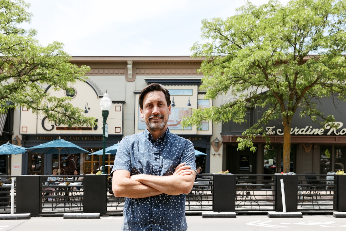An adult male with brown hair stands. with his arms crossed in front of several downtown Plymouth restaurant store fronts.