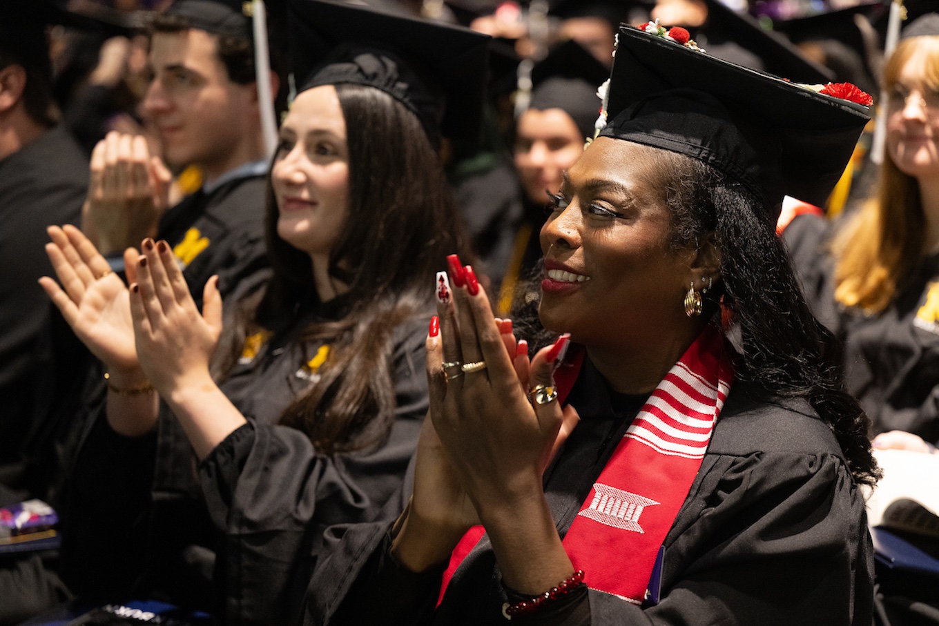 Two smiling women wearing caps and gowns clap as new graduates in the audience
